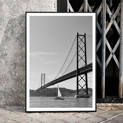 Lisbon Bridge Black and White Poster by Raphael Alves featuring a framed photograph of a suspension bridge and sailboat in the water, framed and displayed in home decor