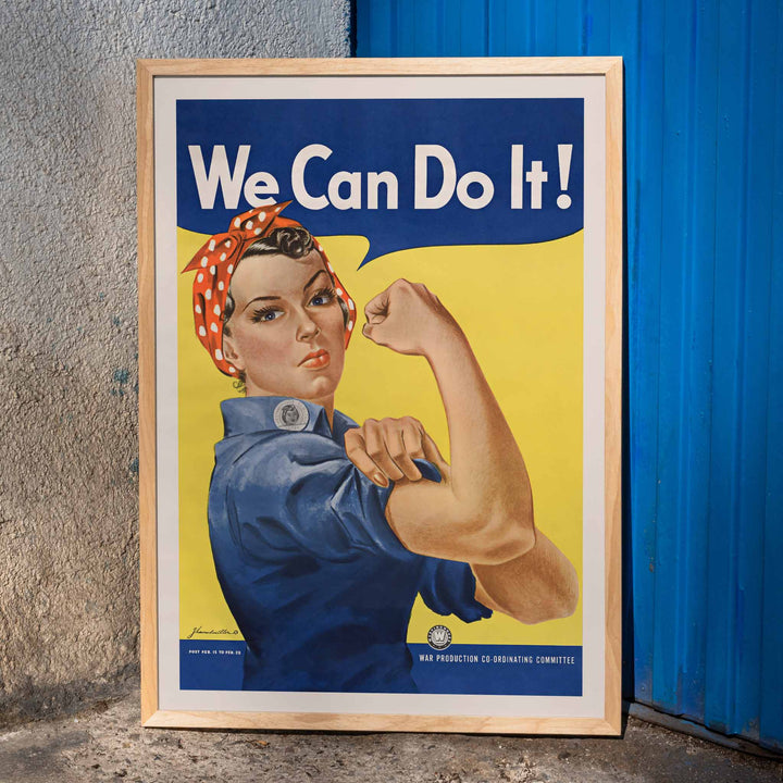 We Can Do It poster by J Howard Miller with determined woman flexing arm in blue and yellow wartime style, framed and displayed in modern living room decor